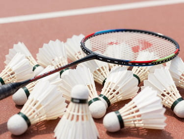 Close-up of a badminton racket resting next to a pile of white shuttlecocks on a court floor, warm natural daylight.