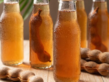 Macro close-up of a chilled condensation-covered glass bottle of tamarind tea next to cracked brown tamarind pods under bright, high-contrast tropical sunlight.