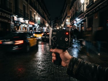 Cinematic low-angle street photography in Madrid at night, motion-blurred lights, a hand holding a vintage video camera with a tiny red tally light glowing, heavy 35mm film grain, moody chiaroscuro