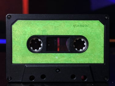 Album cover art showing a close-up of an old audio cassette tape with neon green handwritten labels, dark background.