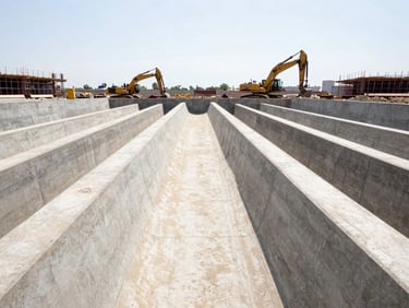 Wide-angle environmental shot of massive concrete drainage channels under construction, heavy machinery moving in the background, bright harsh daylight.