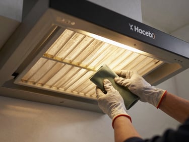 Overhead of a Haceb range hood filter removed and laid on a counter, technician's gloved hands cleaning the grease mesh under warm kitchen tungsten light