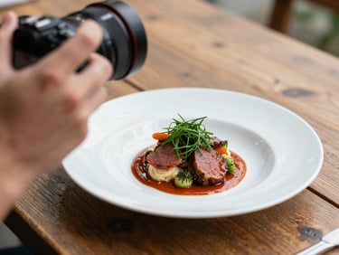 Close-up of a hand holding a camera, shooting a perfectly plated dish on a rustic wooden table, soft natural daylight, shallow depth of field.