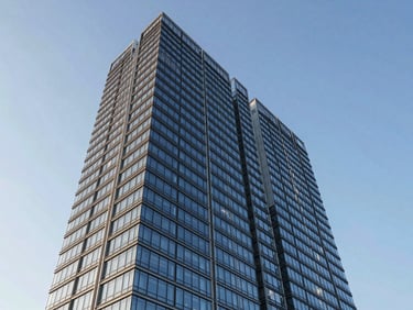 Wide architectural exterior of a contemporary mixed-use tower, clean glass facade catching morning daylight, low-angle framing conveying scale and precision, clear blue sky above, no people