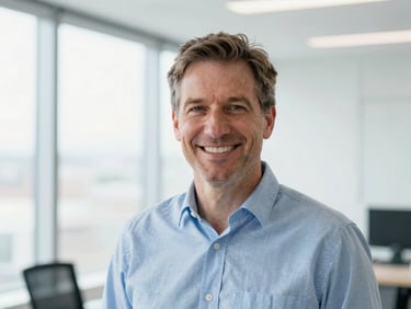 Professional headshot of a distinguished British man, mid-40s, smiling warmly, in a modern, clean office setting, bright natural daylight, clean blue and white tones, shallow depth of field, 35mm.