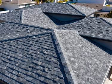 High-angle drone shot of a modern residential roof with dark architectural shingles, crisp lines, under bright California sun, deep blue and slate gray tones, sharp focus, 35mm lens