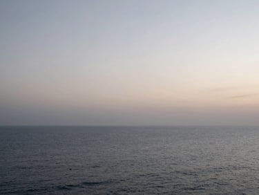 Wide panoramic shot of an ocean horizon at dawn, soft desaturated morning light, overcast sky, vast negative space, calm coastal atmosphere.