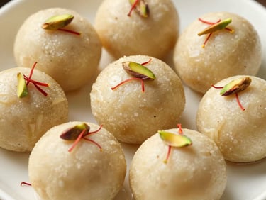 Artisanal Indian sweet pedas arranged on a delicate ceramic dish. Each light beige, round cardamom-infused sweet is topped with a few delicate red saffron strands and a single green pistachio sliver. Soft, warm lighting, elegant and high-end presentation highlighting local dairy heritage.