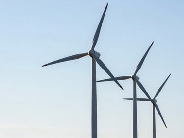 Wide shot of modern wind turbines against a clear sky, showing their scale and integration into the landscape. Industrial editorial photography, bright natural daylight, clean technical details, high contrast, deep slate and steel tones, 35mm.