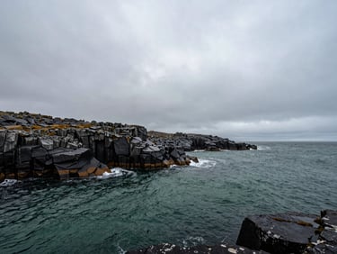 Wide panoramic view of a remote northern coastline under heavy overcast skies, dark basalt rocks, deep green water ripples, misty atmosphere.
