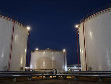 Low-angle dramatic wide shot of massive white liquid storage tanks at dusk, deep navy sky, sharp industrial floodlights reflecting off steel pipelines.