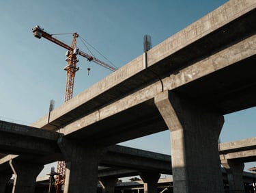 Dramatic low-angle shot of a massive concrete flyover under construction, heavy cranes, steel reinforcement cages visible, intense late-afternoon sun casting long shadows.