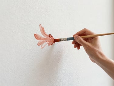 Artist's hands holding a brush loaded with diluted terracotta pigment, painting delicate fluid strokes on a textured white wall, soft natural daylight