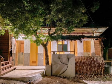 Rustic tropical bungalow at night with bamboo walls, thatched roof, and warm exterior lighting.