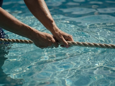 Close-up of hands gripping the pool lane rope at water level, natural sidelight creating caustic light patterns across the water surface, calm water in the foreground