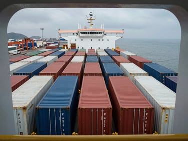 Overhead close-up of a commercial shipping vessel deck showing container rows in tight formation under grey overcast sky, port infrastructure visible at far edge, architectural framing emphasising scale and order