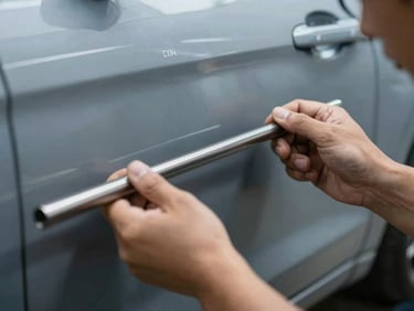 PDR technician's hands gripping a long metal rod inserted behind a car door panel, cool indirect workshop light, slight motion blur on the rod tip showing active pressure being applied, the panel surface visible in background with dent geometry