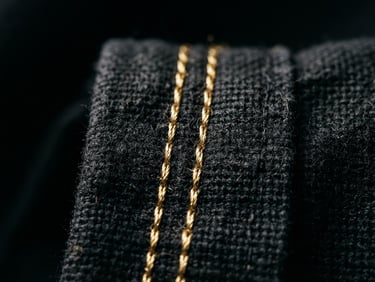 Close-up of precision double-stitch seam on heavy black flag fabric, gold embroidered details catching dramatic high-contrast studio lighting, 35mm photography