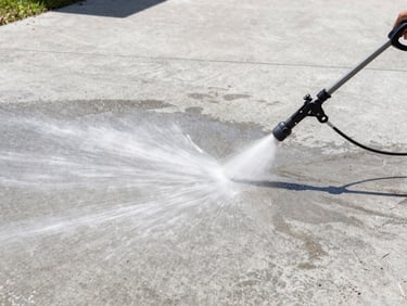 Wide shot of a clean concrete driveway being pressure washed, water spray lifting dirt, bright midday sunlight, sharp focus.