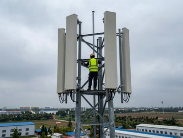 Wide-angle view of a 5G antenna array being mounted on a steel tower, technician in high-visibility vest at height, overcast sky, industrial landscape below