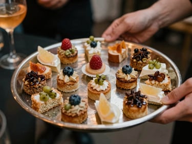 Close-up overhead of cocktail hour passed hors d'oeuvres on a round silver tray, bite-sized items neatly arranged, warm ambient lighting, staff hands visible at tray edges