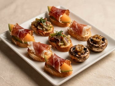 Overhead close-up of a white rectangular serving platter with six neatly arranged appetizer bites — prosciutto-wrapped melon, bruschetta, and stuffed mushrooms — warm even studio lighting on a cream linen surface