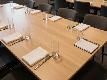 Wide overhead view of a corporate luncheon table fully set — rectangular tables, water glasses aligned, folded napkins, notepads at each place, warm even office-venue lighting, empty before attendees arrive