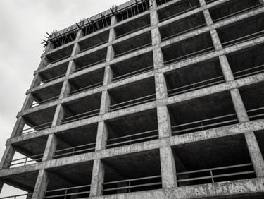 Wide architectural shot looking up the facade of a concrete mid-rise under construction in an urban Pacific Northwest setting, steel rebar grid visible through open floor plates, overcast diffused daylight, stark high-contrast monochrome
