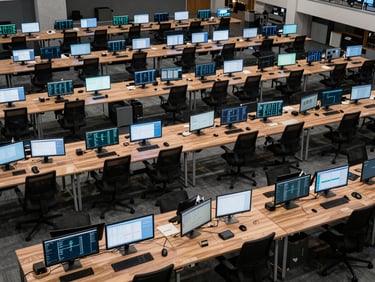 Wide architectural interior shot, overhead perspective looking down at a structured grid of desks with monitors displaying data dashboards, cool studio lighting casting sharp shadows, no people visible, emphasis on geometric order and operational infrastructure