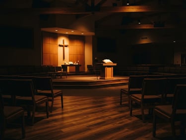 Warm moody photography of an empty modern church sanctuary, low-key warm amber stage lighting casting long soft shadows on wooden panels, intimate peaceful atmosphere, shallow depth of field