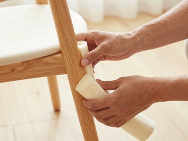 Close-up of professional hands wrapping a wooden chair leg with thick protective foam, warm natural light.