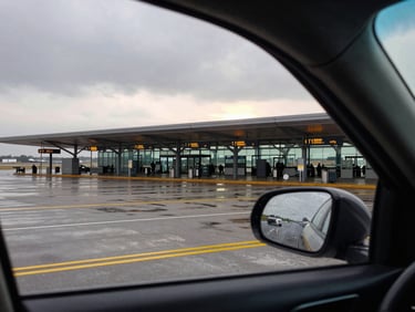 Close framing through a taxi windshield of an airport terminal entrance at dawn, overcast sky, yellow taxi lane markings visible on wet tarmac, departure signs just legible in background
