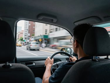 Passenger perspective from rear seat of a taxi, city buildings scrolling past the side window under grey midday light, driver's headrest and hands on wheel visible in foreground