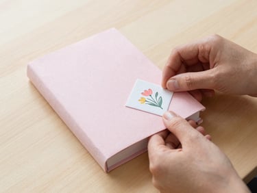 Hands carefully wrapping a pastel notebook in pink tissue paper, placing a custom floral stamp on the box, soft morning daylight, cozy flatlay