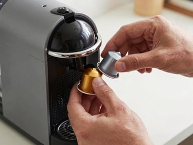 Technician hands inserting a Nespresso capsule module back into machine casing, close-up precision grip, bright north-facing daylight