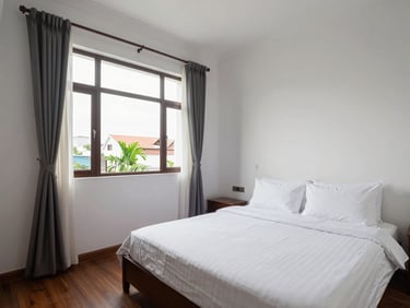Interior bedroom of a furnished apartment in Thanh Khê district Đà Nẵng, natural window light, queen bed with clean linen, white walls, wide framing showing full room context