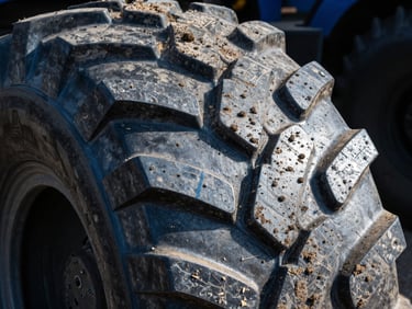 Detailed macro shot of a massive tractor tire tread caked with dry earth, high-contrast daylight highlighting the deep rugged pattern.