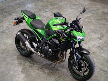 Overhead three-quarter view of a Kawasaki Z900 on showroom concrete floor, green and black bodywork under hard strobe light, instrument cluster visible, no rider