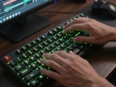 Extreme close-up overhead shot of hands on a mechanical keyboard, monitor glow reflecting neon-green on fingertips, dark desk surface, editing software visible blurred in background