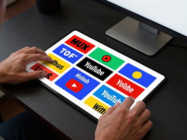 Overhead shot of hands arranging YouTube thumbnail mockups on a dark desk, high-key monitor light illuminating bold typography and saturated color-blocked graphic layouts
