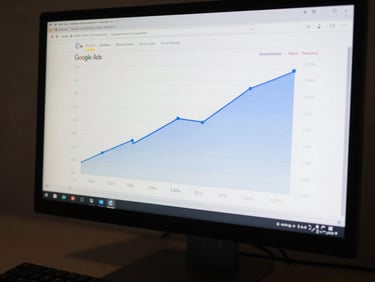 Close-up of a monitor displaying Google Ads campaign performance graph with upward inflection, cool studio key light, keyboard partially visible at bottom, no people