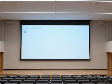 Wide architectural shot of a contemporary campus auditorium, large high-resolution presentation screen active, sleek acoustic paneling, clean rows of seating, bright natural daylight mixing with subtle screen glow.