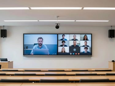 Wide architectural shot of a university lecture hall, showing a dual-display setup at the front with remote students visible on one screen, clean ceiling-mounted beamforming microphones, bright daylight.