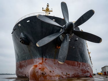 Extreme wide-angle shot of a bulk carrier in a drydock, exposing the massive hull and propeller blades under overcast daylight.