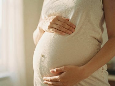 Pregnant woman in soft natural light, hands on belly, warm editorial photography, cream and earth tones, shallow depth of field