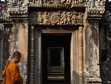 Intimate close-up of a Cambodian temple doorway at dawn, stone carvings in soft raking morning light, a monk's saffron robe partially visible at the left edge, no tourists, no flash