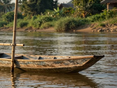 Close-up environmental frame, golden-hour light catching the surface of a calm river in Luang Prabang, a narrow wooden boat tied to a bamboo pole in the foreground, lush green riverbank blurred behind, natural warm afternoon light