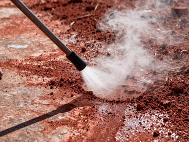 Close-up of a power washing wand blasting clean a path through dark red dirt on concrete, steam rising, high-contrast sunlight, 35mm