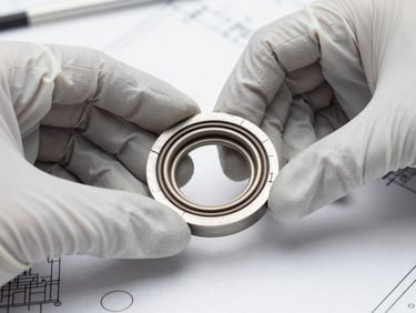 Hands in safety gloves holding a failed mechanical seal face under bright studio lighting, cracked seal ring and scored face visible in sharp macro detail, clean white inspection surface underneath, technical blueprint partially visible at the frame edge