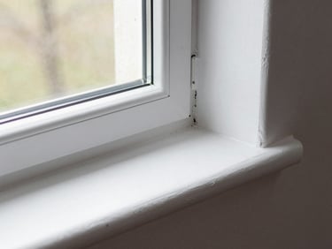 Close-up angled shot of a window casing and sill freshly painted in bright white against a freshly painted wall, showing the clean masking line at the glass edge with zero bleed, natural daylight from the window itself lighting the detail